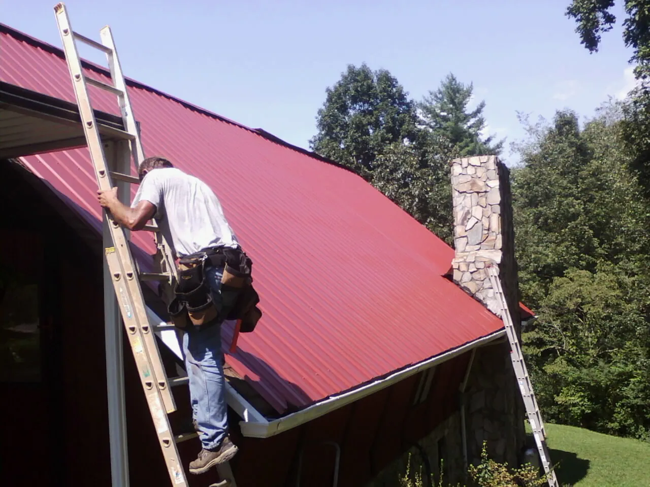 Roofer on a ladder beside a red metal roof and stone chimney — Soffit & Fascia Repair by Roofing Brothers in Eldon
